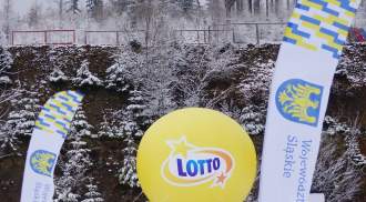 Żółty balon z napisem Lotto, strefa startu biegu narciarskiego z dwiema chorągiewkami  Województwa Śląskiego, w tle zimowa sceneria ze świerkami i porośnięte zbocze.