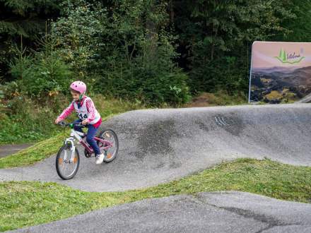 Mała dziewczynka w różowym kasku i jasnej bluzie z numerem startowym jedzie na rowerze po torze pumptrack. W tle widać baner reklamowy z napisem Istebna oraz gęsty, zielony las.
