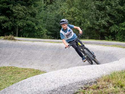 Chłopiec w kasku i niebieskiej koszulce z numerem 205 dynamicznie pokonuje zakręt na rowerze górskim po betonowym torze pumptrack. W tle gęsty las, na pierwszym planie krawędź trawy.