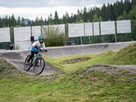 Chłopiec w kasku i jasnej koszulce jedzie na rowerze górskim po profilowanym torze pumptrack. Otaczają go zielone, trawiaste pagórki, a w tle widać ogrodzenie oraz gęsty las pod pochmurnym niebem.