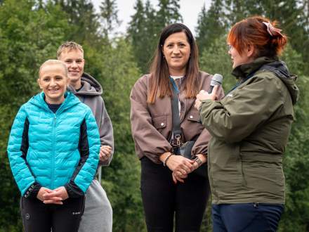 Kobieta w zielonej kurtce przemawia do mikrofonu podczas wydarzenia na świeżym powietrzu. Obok niej stoją uśmiechnięci uczestnicy: dwie kobiety oraz młody mężczyzna. W tle widać gęste, zielone drzewa.
